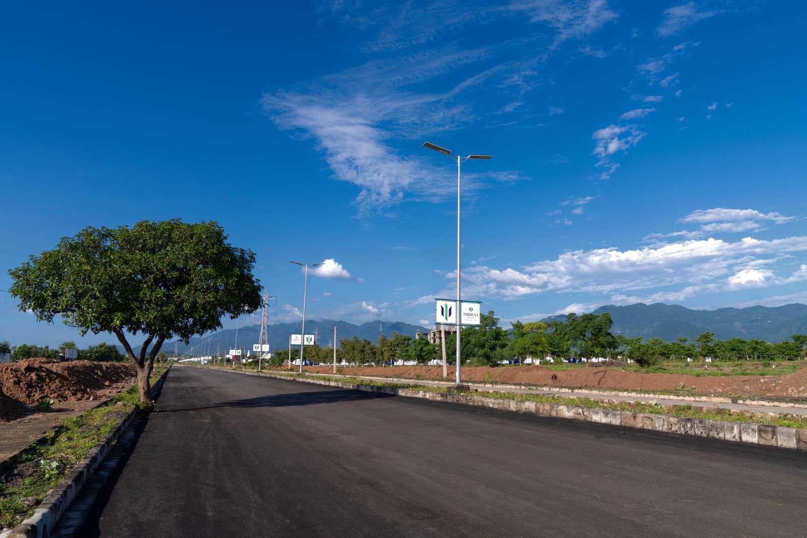 Photograph of a wide new road with solar street lights and Trident signs inside Trident Hills, Pinjore, with the hills behind