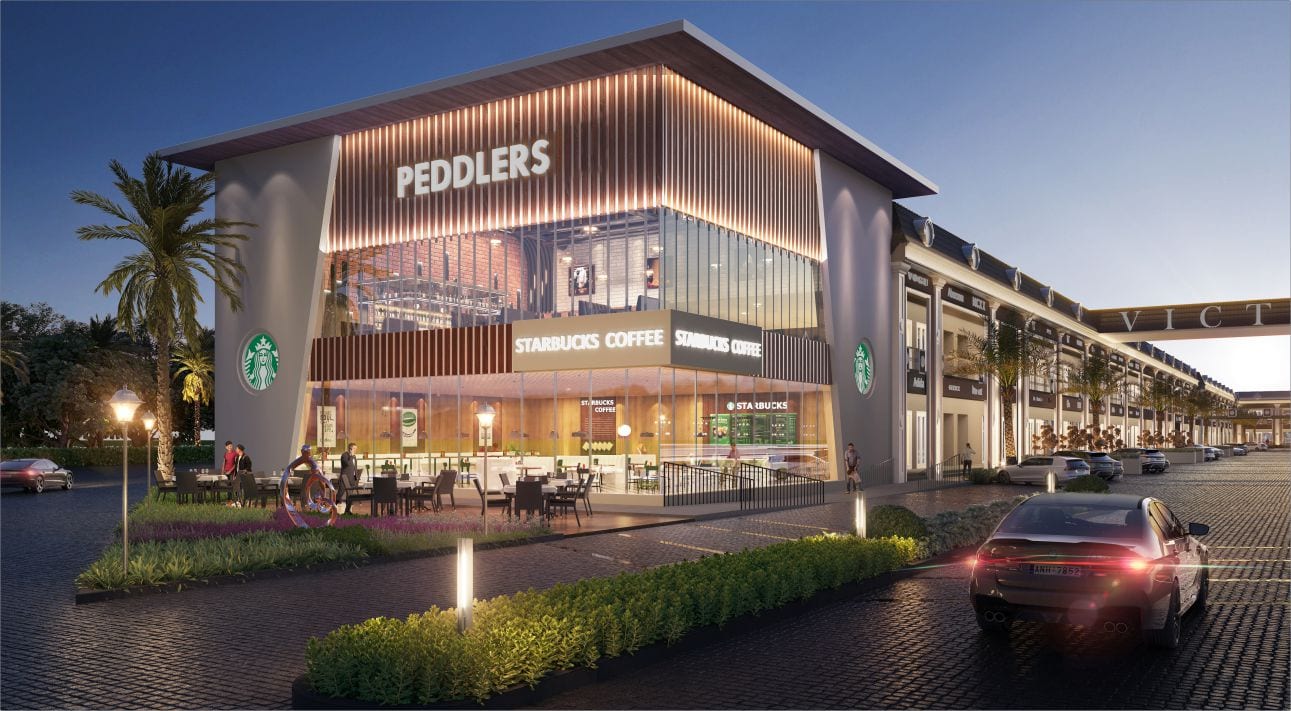 Evening view of the glass-fronted corner cafe and restaurant block with outdoor seating at Victoria Street, Sirhind, with the showroom row and gateway behind.